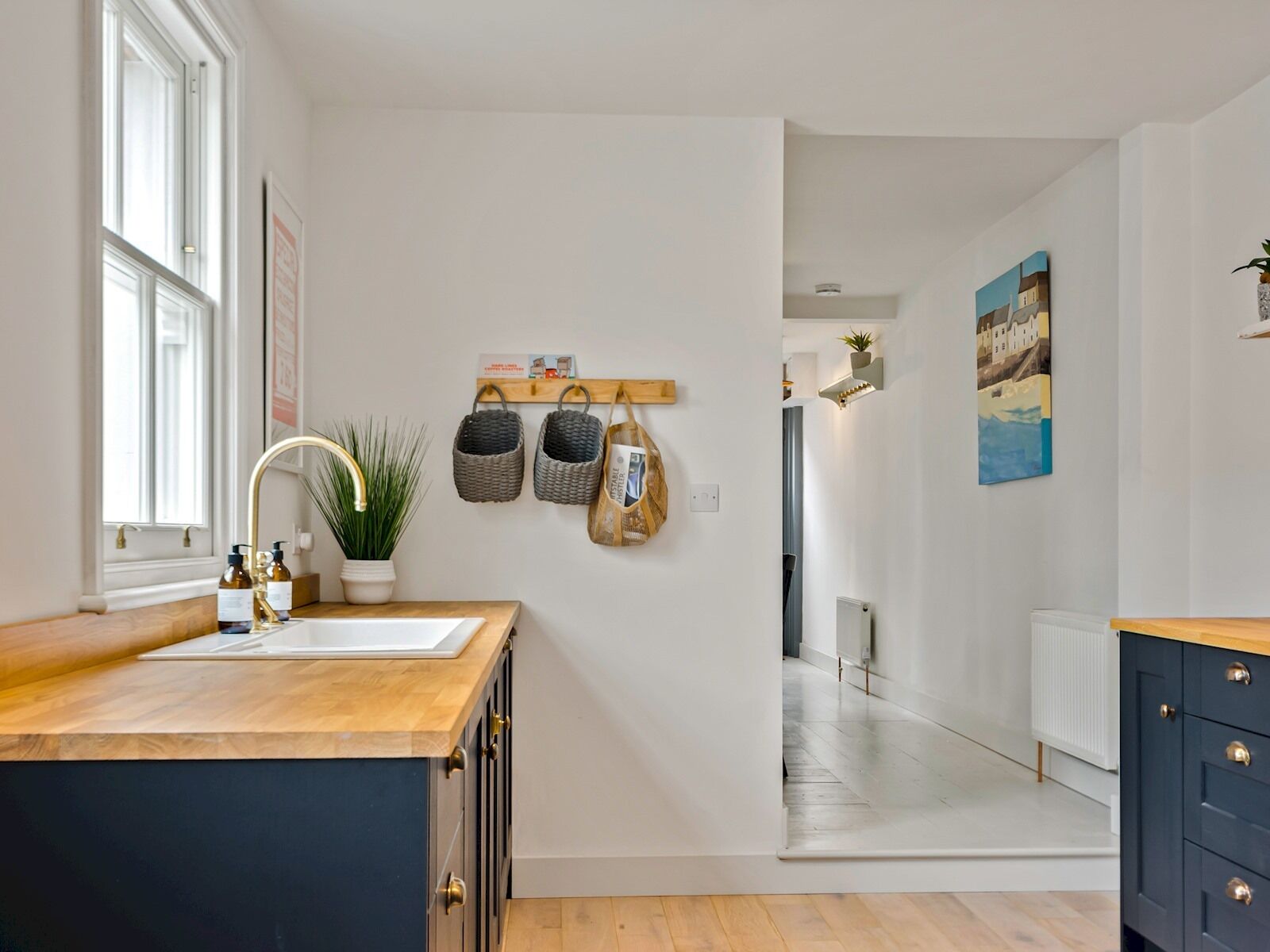 A modern kitchen with a wooden countertop, a sink, and a potted plant. There are baskets hanging on the wall and a painting above the cabinets.