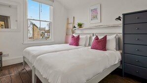 Un dormitorio con un ventanal grande, una cama con ropa blanca y almohadas rosas, un aparador y una pequeña planta sobre él.