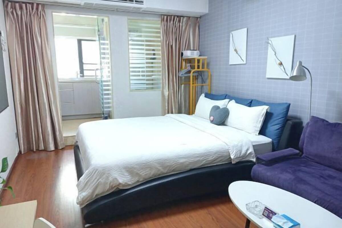 Un dormitorio con una cama, un sofá morado, una mesa blanca y un ventanal con cortinas.