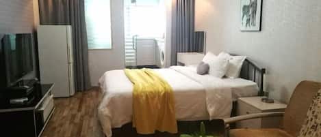 A hotel room with a bed, a chair, a TV, a refrigerator, a washing machine, and a plant.