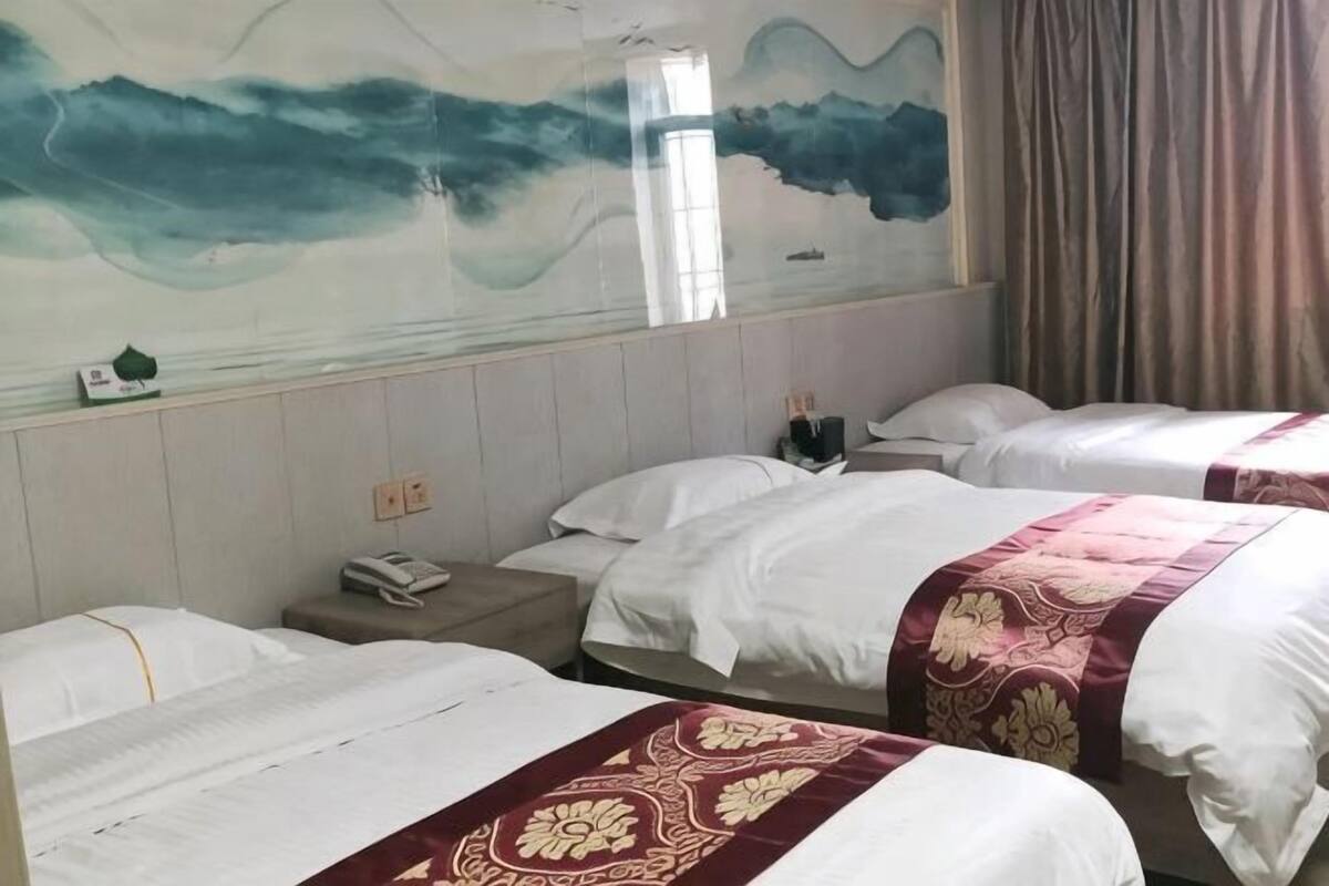 Hotellihuone, jossa on kolme sänkyä, seinällä suuri seinämaalaus, yöpöydällä puhelin ja ikkunassa verhot.
