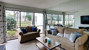 Amplia sala de estar con dos sofás, una mesa de centro y un televisor. Ventanas grandes ofrecen vistas al exterior.