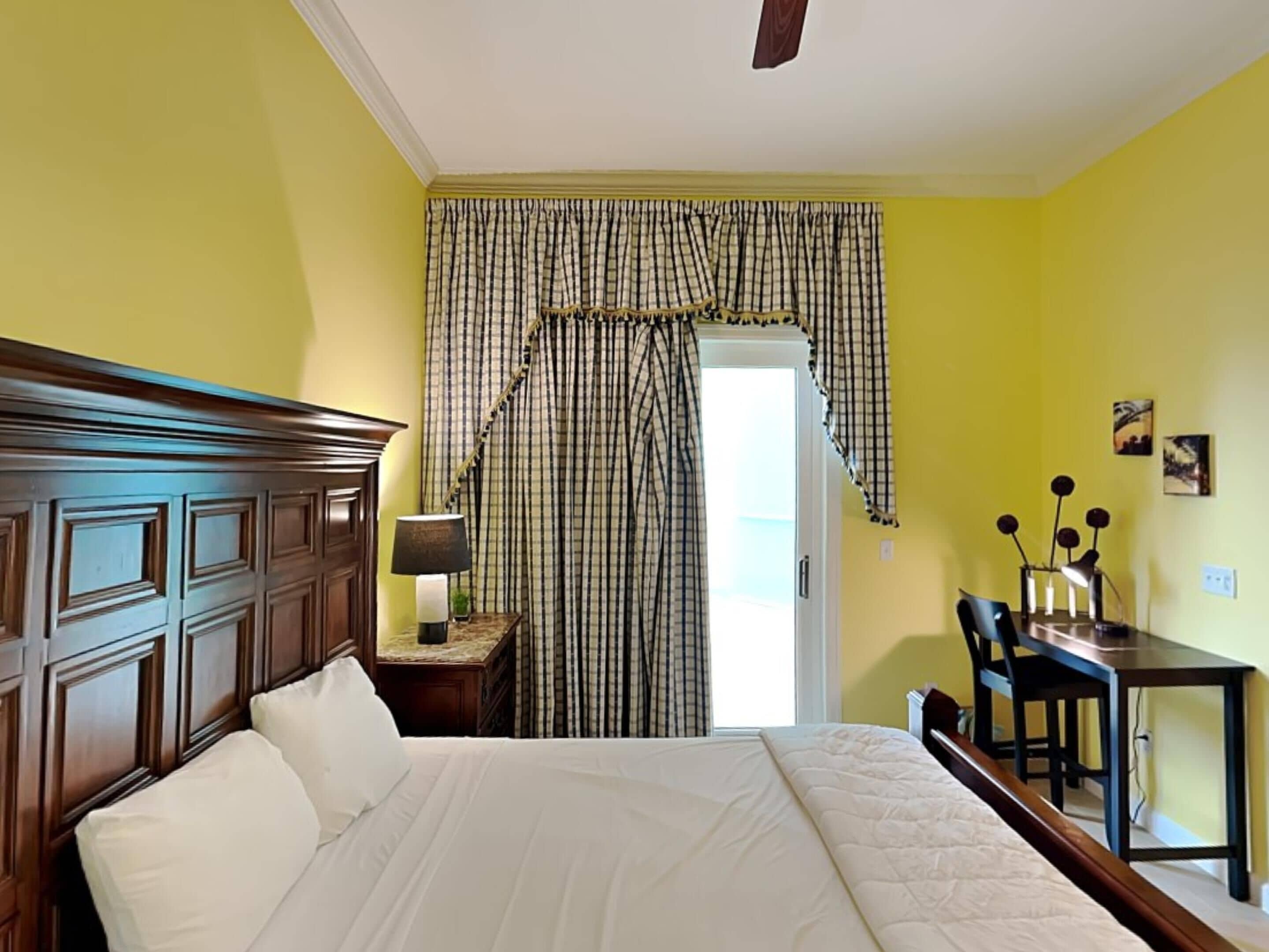 Una camera da letto con una testiera in legno, un letto con lenzuola bianche, un comodino con una lampada, una tenda a quadri e una scrivania con sedie.