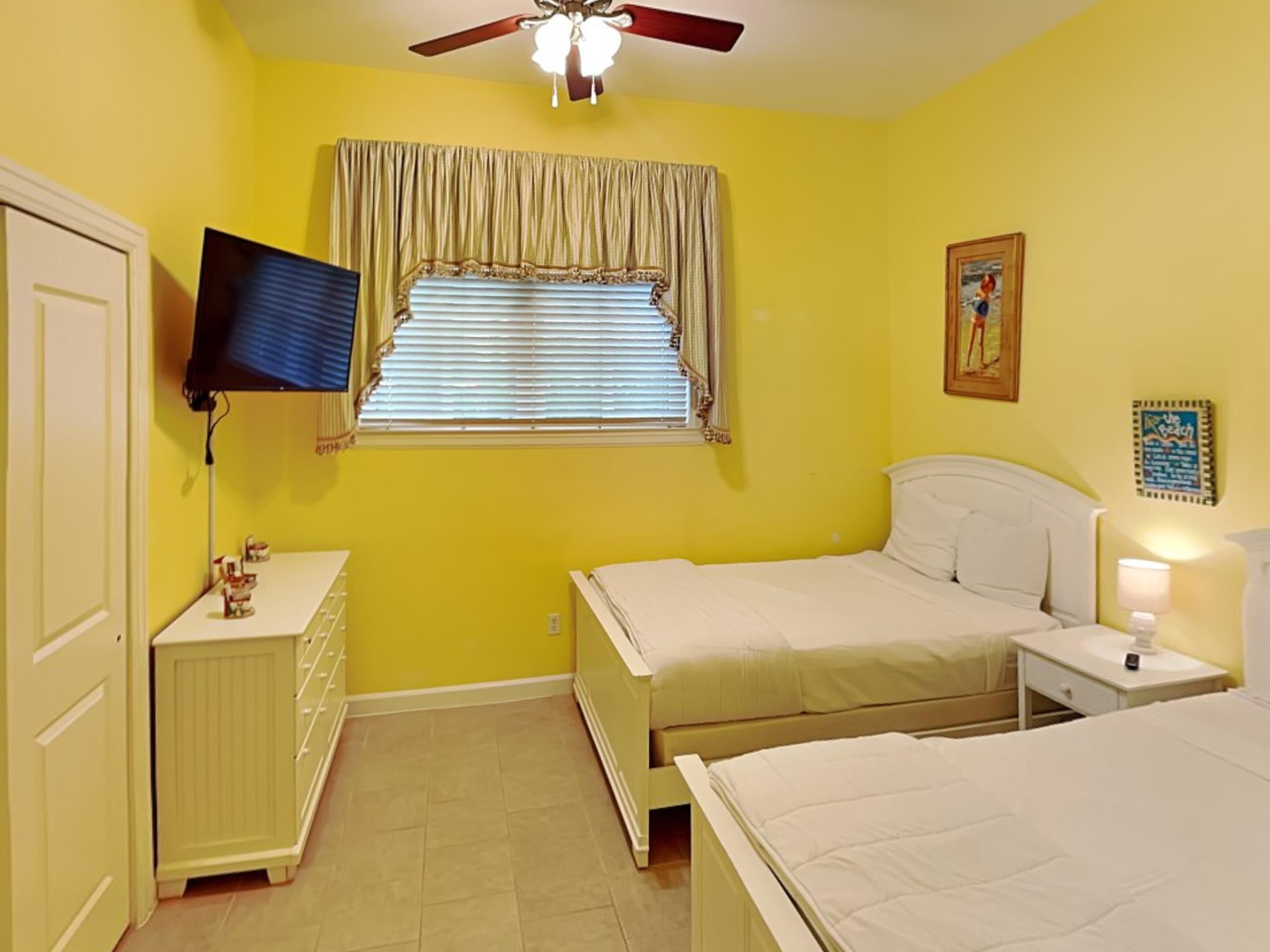 A bedroom with two beds, a ceiling fan, a TV mounted on the wall, and a window with curtains.