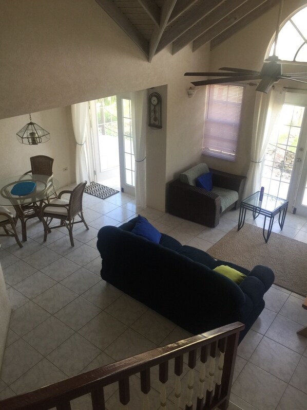 A spacious living area with a large dark-colored sofa, a dining table with wicker chairs, a ceiling fan, and a view through a glass door.