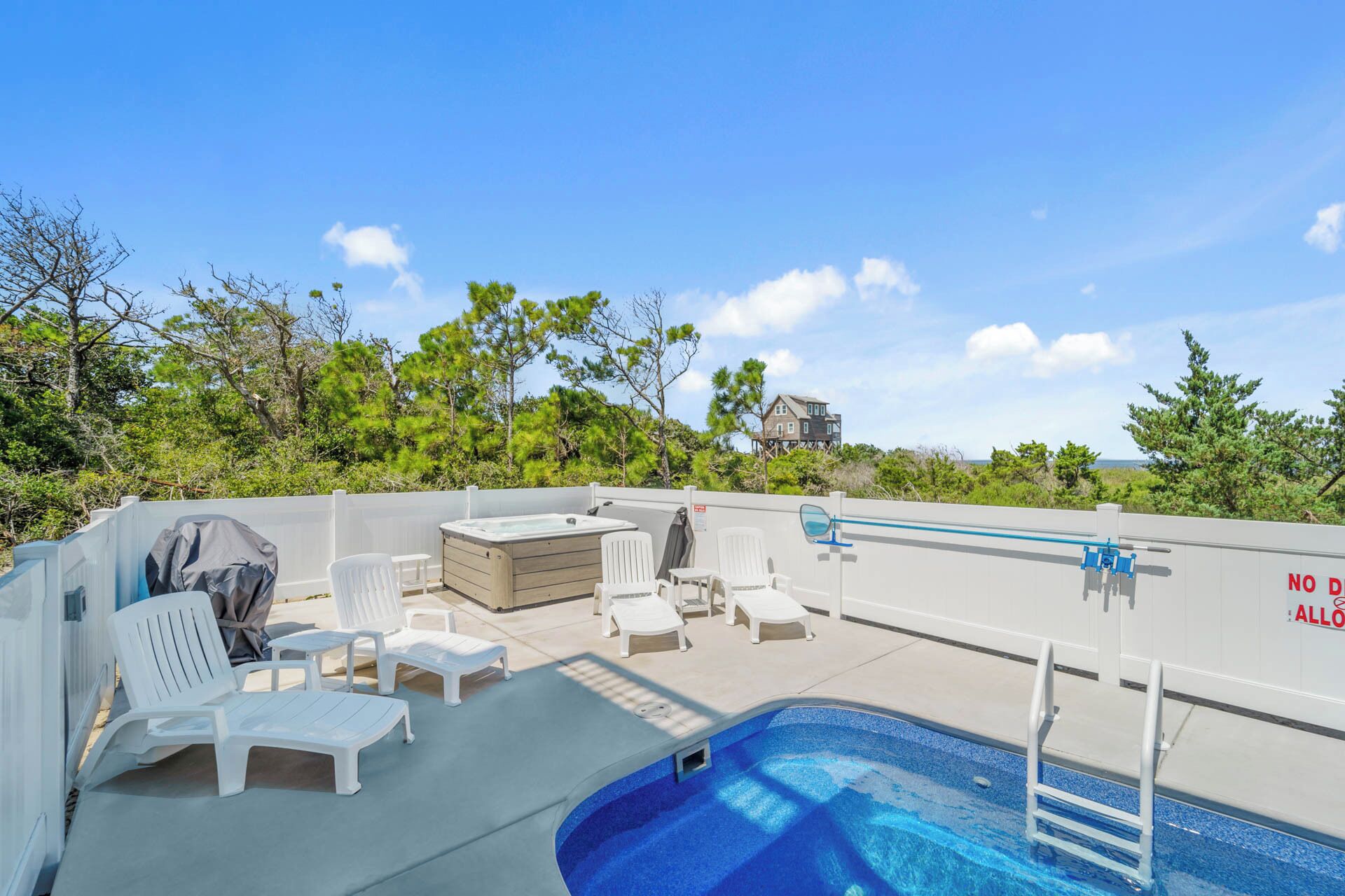 A rooftop pool area with lounge chairs and a hot tub, surrounded by trees and a view of a house.