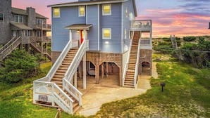 A multi-story house with a prominent staircase leading to the entrance, surrounded by lush greenery.