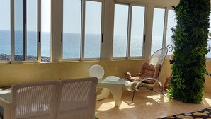 A room with a view of the sea, a wicker chair, a small table, and a green vertical garden.