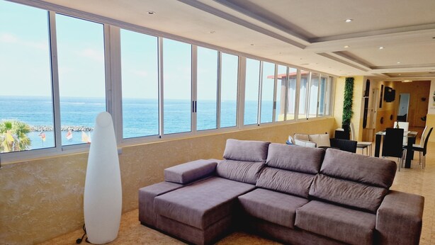 A modern living room with a large sectional sofa, a white surfboard, and a view of the ocean through large windows.