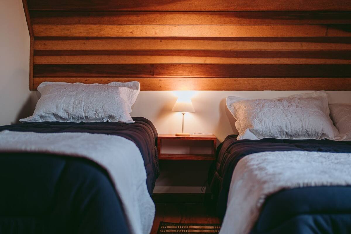 A room with two beds, each with a white pillow and a dark blanket. There is a wooden headboard, a nightstand with a lamp, and a small table with a keyboard.