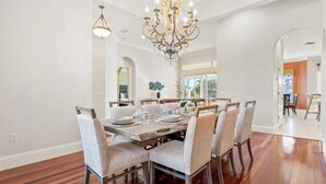 Une salle à manger avec une table en bois, des chaises rembourrées, un lustre et une vue sur l’extérieur.