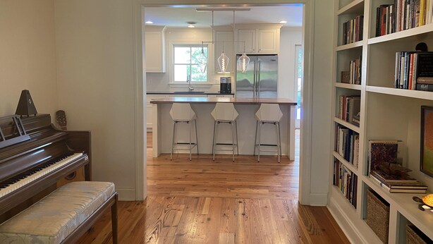 A spacious living room with a piano, a bookshelf, and a kitchen area in the background.