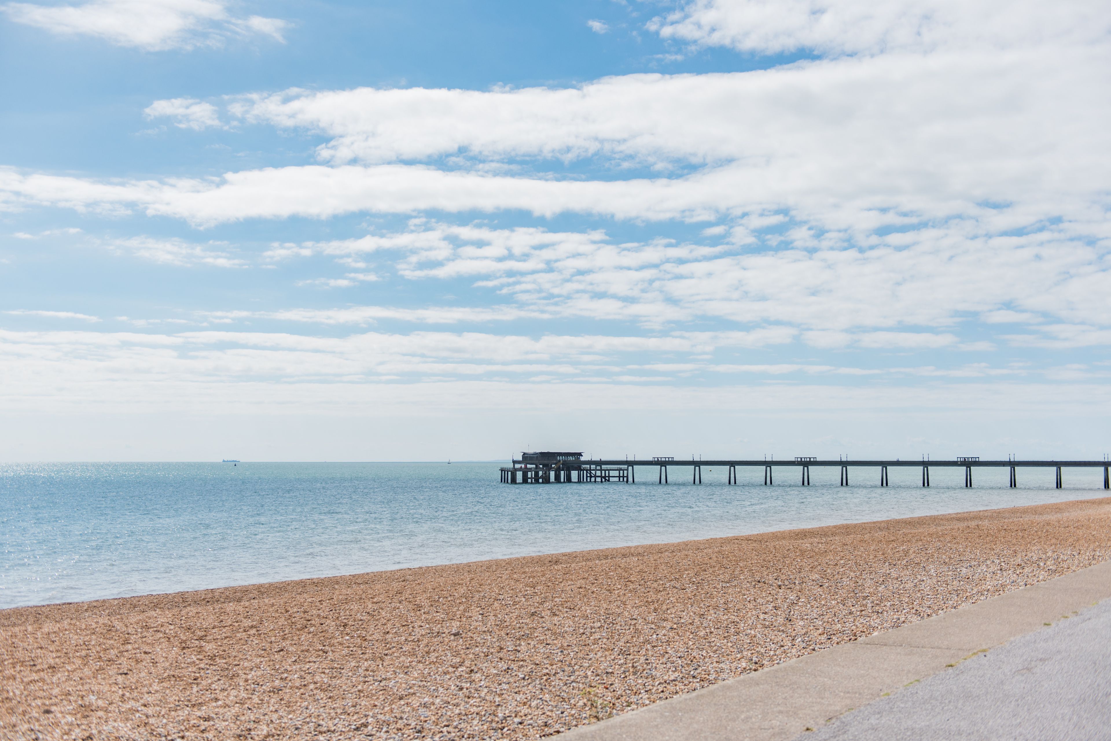 A pebbled beach with a pier extending into calm waters under a partly cloudy sky.