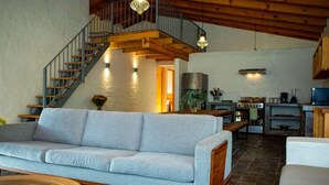A spacious living area with a light grey sofa, a wooden coffee table, a staircase with a wooden railing, and a kitchen area with a stove and a sink. - Cabañas La Serenisima (Tapalpa)