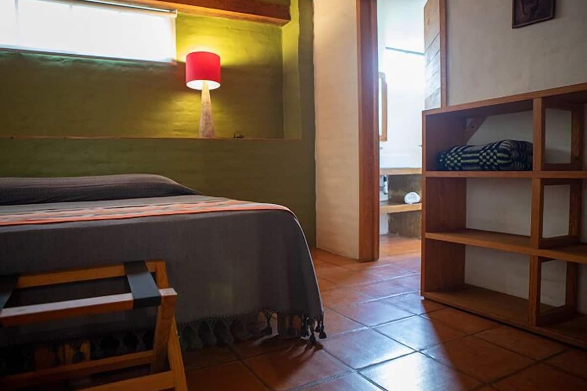 A bedroom with a bed, a wooden bench, a red lamp, and a shelf with folded clothes.