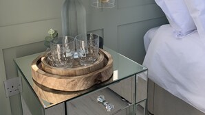 Une chambre moderne avec une table de chevet en verre, un plateau en bois contenant des verres en cristal, une bouteille en verre transparent et une lampe noire.