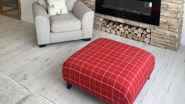 Un salon avec une cheminée en pierre, un canapé gris orné d’un coussin à motifs de cerfs et un pouf rouge à carreaux.