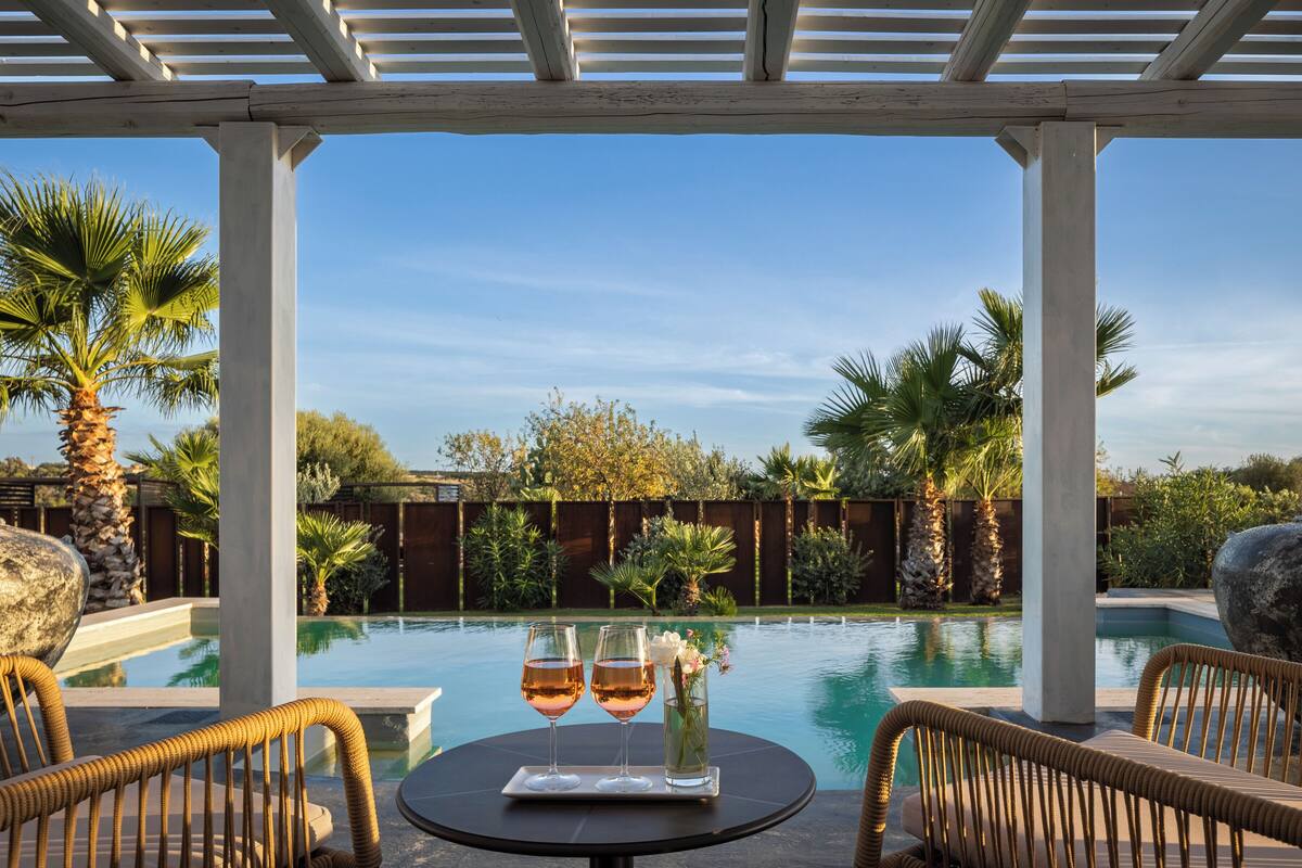 Un espace aménagé au bord de la piscine, avec des fauteuils en osier, une table sur laquelle reposent deux verres de vin et un vase rempli de fleurs.
