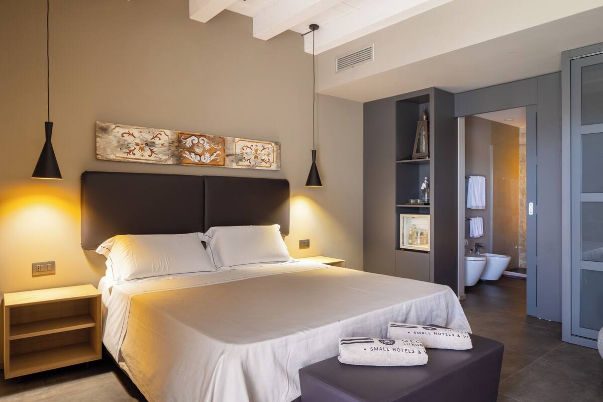 Une chambre d’hôtel moderne avec un grand lit, des lampes de chevet, une table de chevet, une petite table avec des serviettes et une salle de bain visible à travers une porte ouverte.
