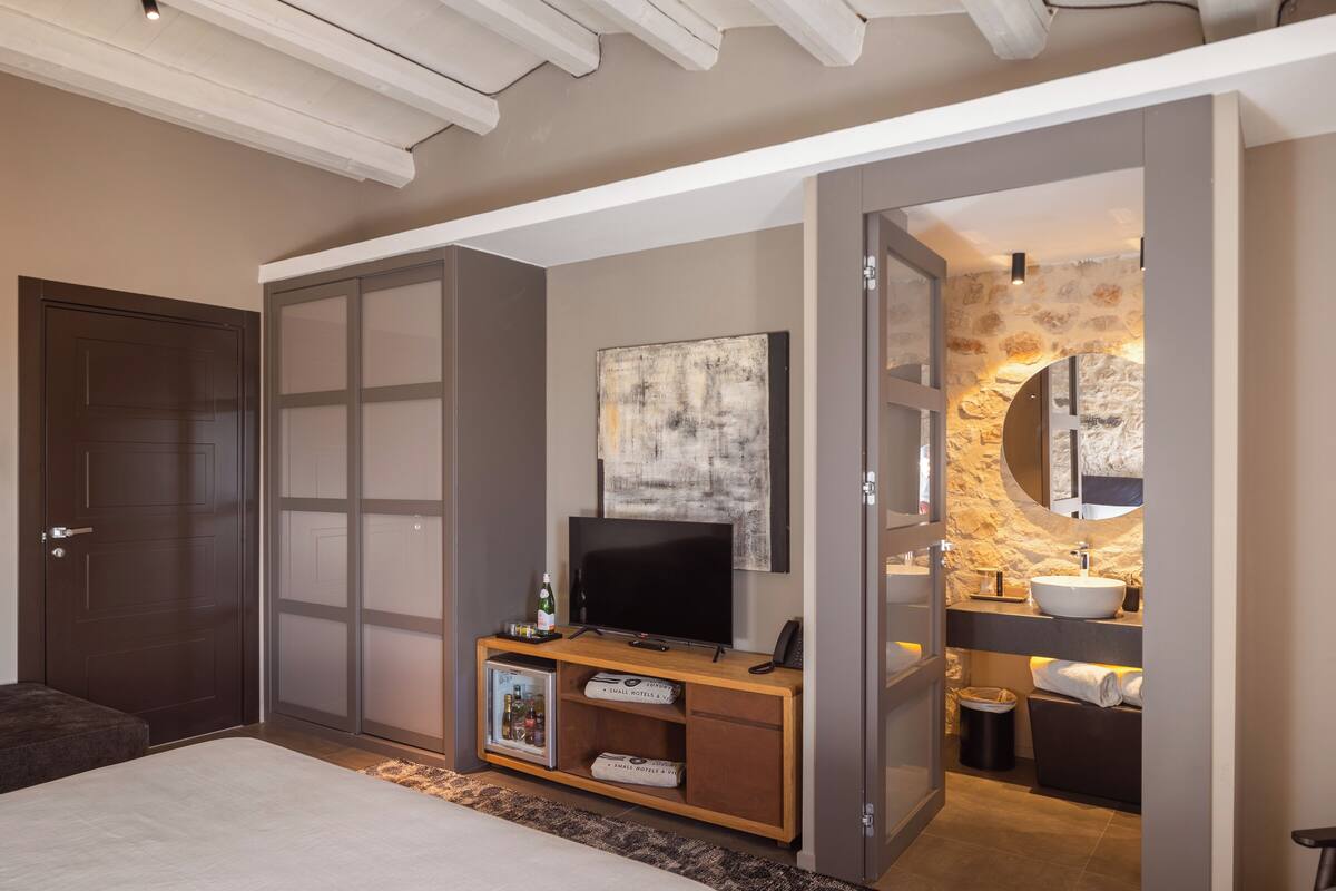 Une chambre moderne avec un grand lit, un téléviseur posé sur une console en bois, une salle de bain avec un mur en pierre et une porte donnant sur une autre pièce.