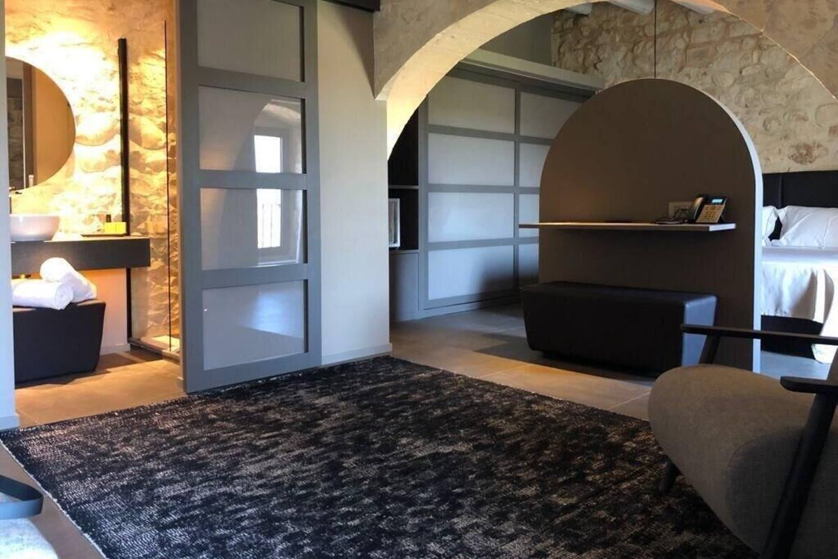 Une chambre d’hôtel moderne avec un mur en pierre, un grand miroir, un lavabo rond, un canapé et un coin lecture avec une étagère.
