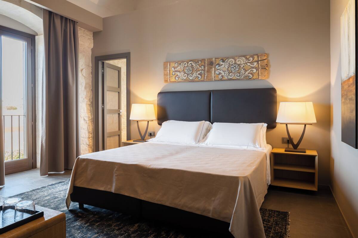 Une chambre d’hôtel avec un grand lit, deux lampes de chevet, un élément décoratif mural, une petite table d’appoint et une vue sur l’extérieur grâce à une porte coulissante.