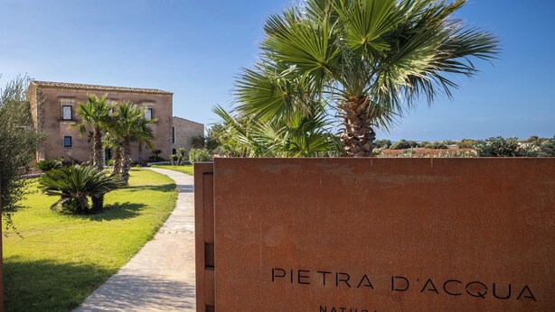Schild für Pietra d'Acqua Natural Retreat Spa Resort, mit einem Weg zu einem von Palmen umgebenen Gebäude.