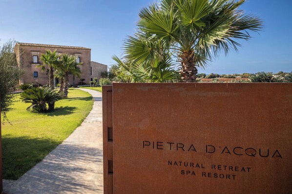 Schild für Pietra d'Acqua Natural Retreat Spa Resort, mit einem Weg zu einem von Palmen umgebenen Gebäude.