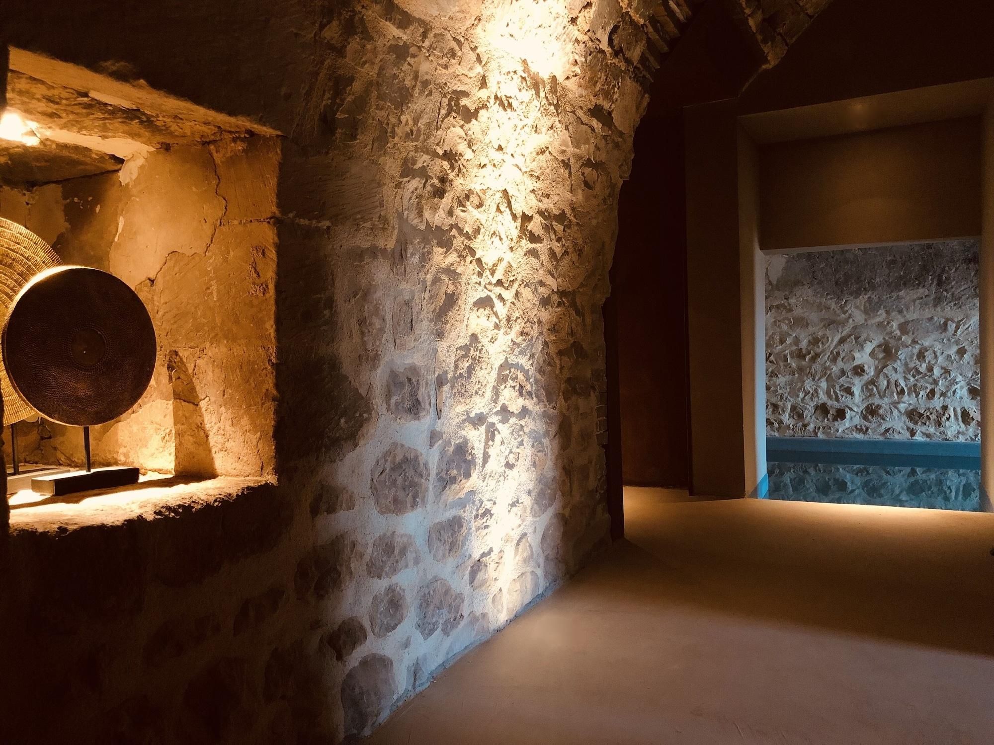 Ein schlicht beleuchteter Raum mit rauen Steinwänden, einem runden Tisch mit Lampe und einer gemusterten Wand im Hintergrund.