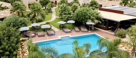 A pool area with lounge chairs and umbrellas, surrounded by palm trees and a stone building.