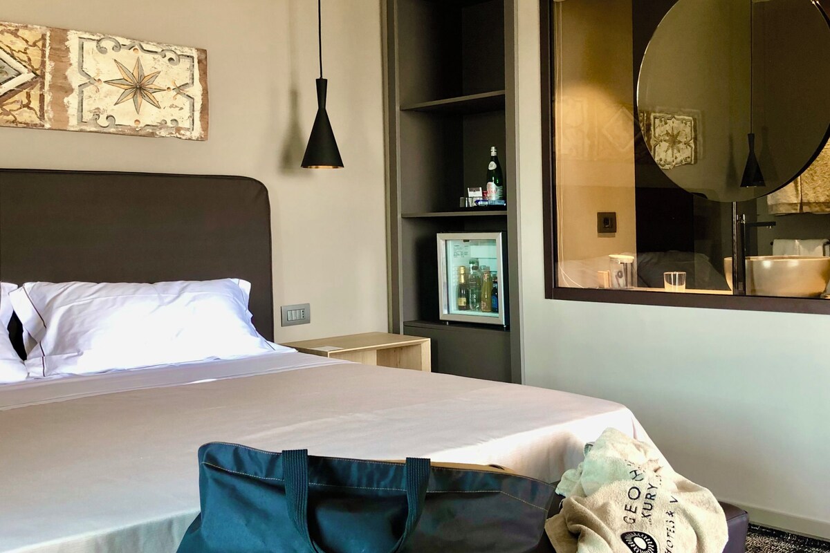 Ein Hotelzimmer mit Bett, Nachttisch, Minibar, großem Spiegel und Wand mit dekorativen Fliesen.