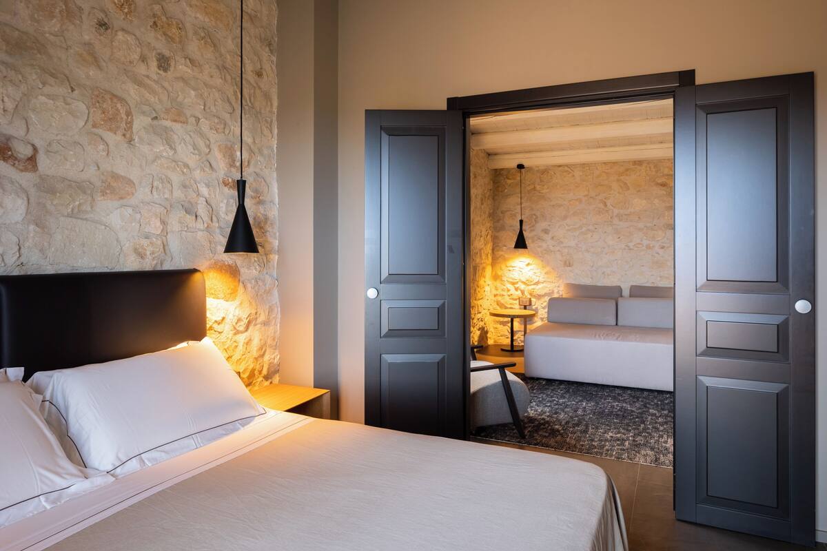 Une chambre d’hôtel avec un mur en pierre, un lit avec du linge de maison blanc, une table de chevet, une lampe et un canapé dans la pièce adjacente.