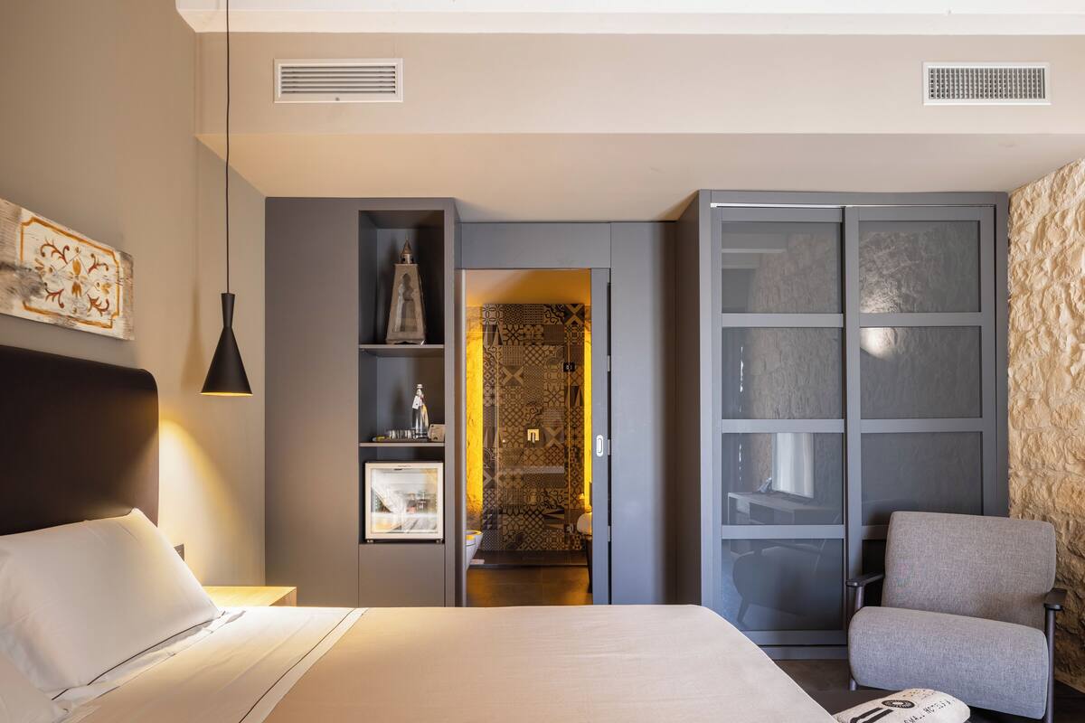 Une chambre à coucher moderne, avec un lit, une chaise, une armoire et une salle de bain visible à travers une porte ouverte.