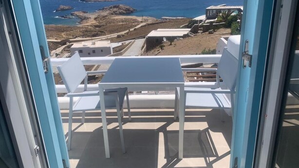 Un balcón con mesa y sillas, con vistas a un paisaje costero con afloramientos rocosos y mar azul cristalino.
