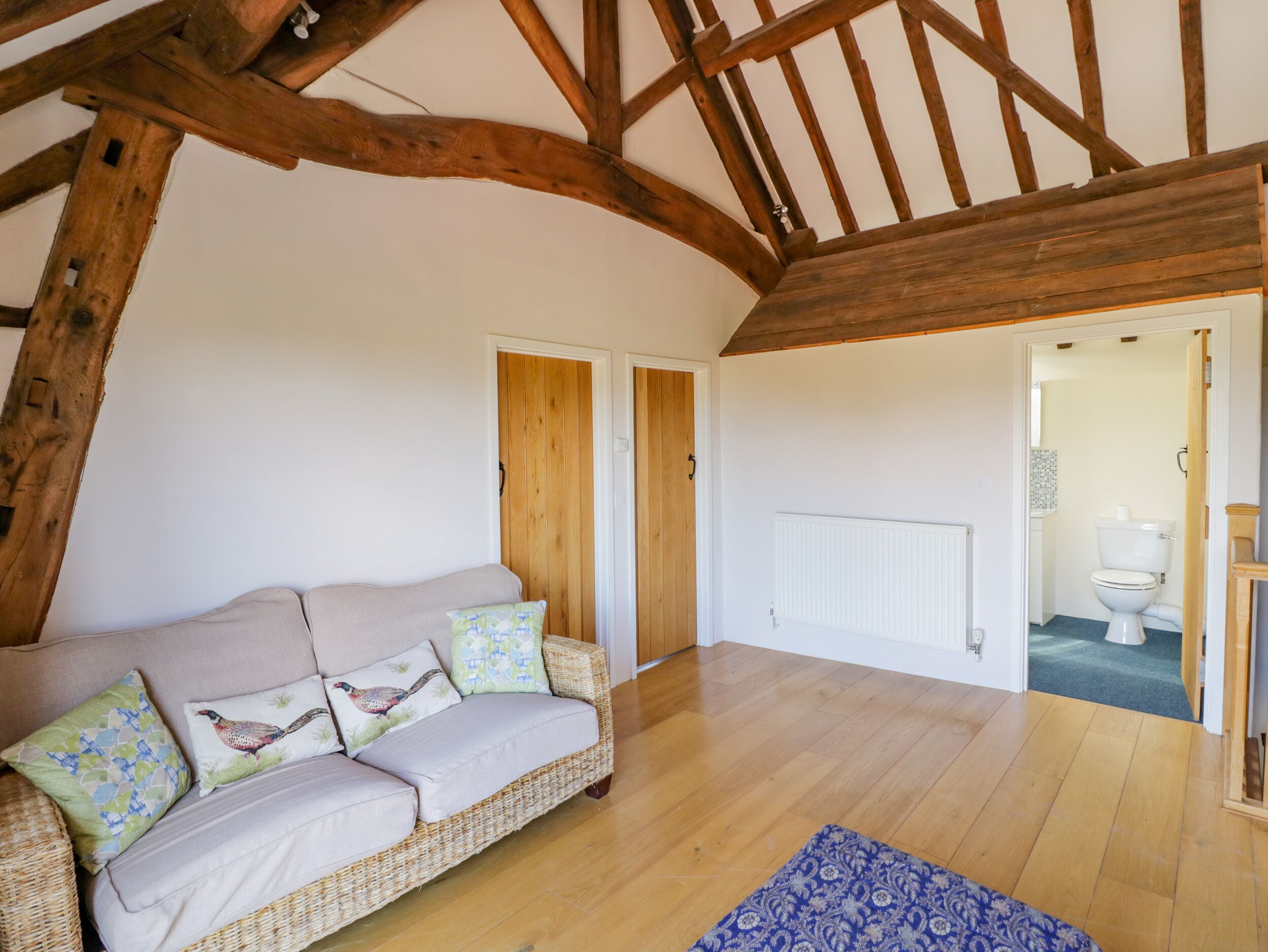Un salon chaleureux avec un plafond haut et des poutres apparentes, un canapé beige agrémenté de coussins à motifs et un tapis bleu à motifs posé sur le plancher en bois.