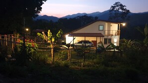 Una casa de dos pisos con un automóvil estacionado frente a ella, rodeada de follaje y montañas al fondo.