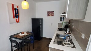 Una cocina compacta con un refrigerador negro, una mesa de comedor con sillas, una lámpara colgante y un lavamanos pequeño.