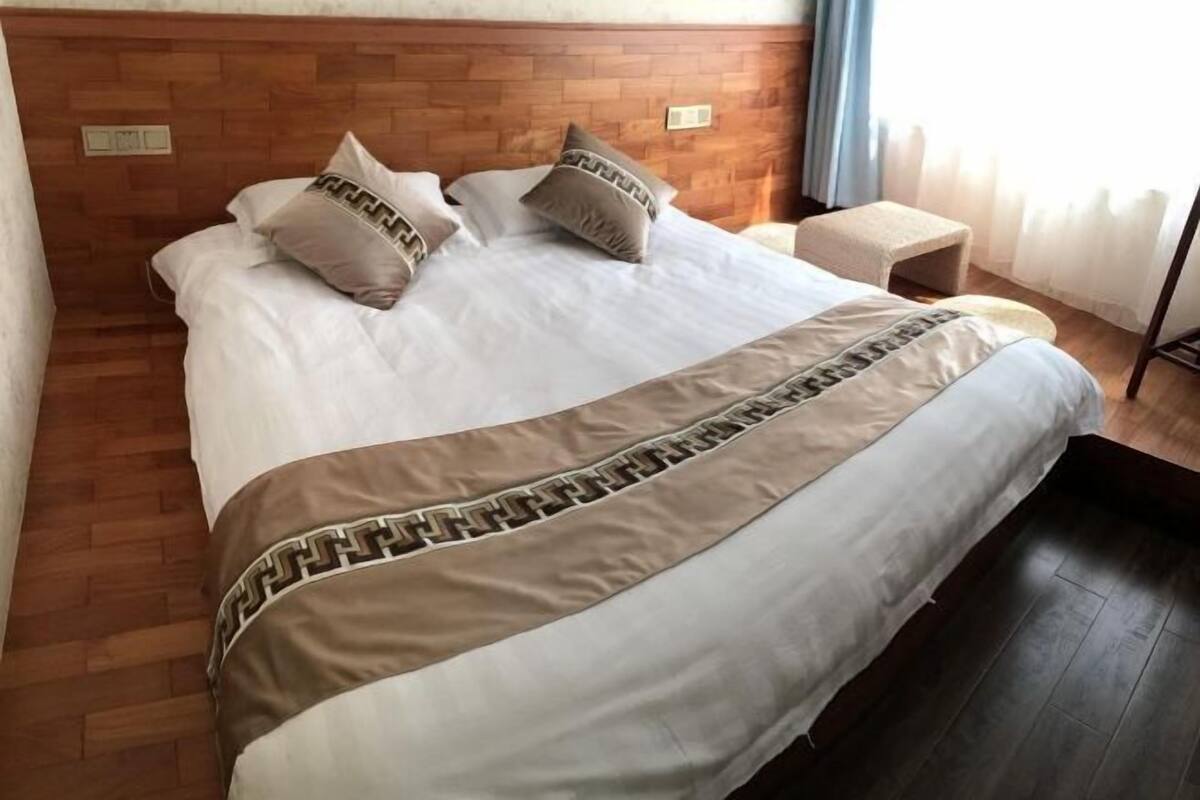 Ein Hotelzimmer mit einem hölzernen Kopfteil, einem Bett mit weißer Bettwäsche und gemusterten Kissen, einem kleinen Nachttisch und einem Fenster mit Vorhängen.