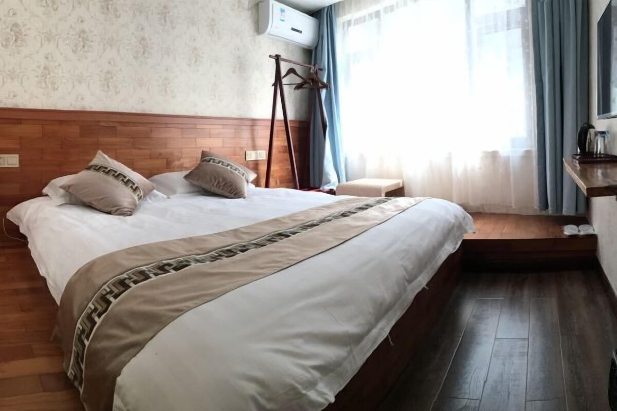 Habitación de hotel con cabecera de madera, una cama grande con colcha color beige y marrón, piso de madera y una ventana con cortinas azules.