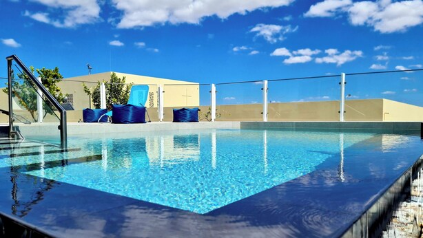 Une piscine à l’eau d’un bleu limpide, entourée d’un petit muret et d’une rambarde métallique. On y trouve deux chaises longues bleues et une petite table avec un coussin bleu. En arrière-plan, on aperçoit un bâtiment doté d’un toit plat et un ciel bleu clair parsemé de quelques nuages.