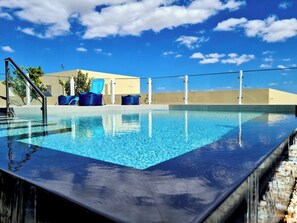 A swimming pool with clear blue water, surrounded by a low wall and a metal railing. There are two blue lounge chairs and a small table with a blue cushion. The background features a building with a flat roof and a clear blue sky with scattered clouds. - Hostel Supremo (Silves)