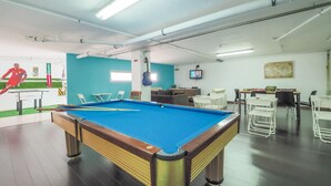 Une table de billard dans une pièce aux murs bleus, au plafond blanc, et où est accrochée une télévision.