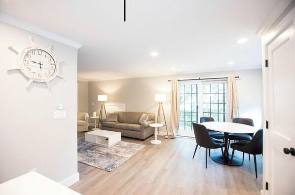 A modern living room with a sofa, coffee table, and dining table with chairs. There is a large window with curtains, a clock on the wall, and a door on the right. - Mid Island Townhouse (Nantucket)