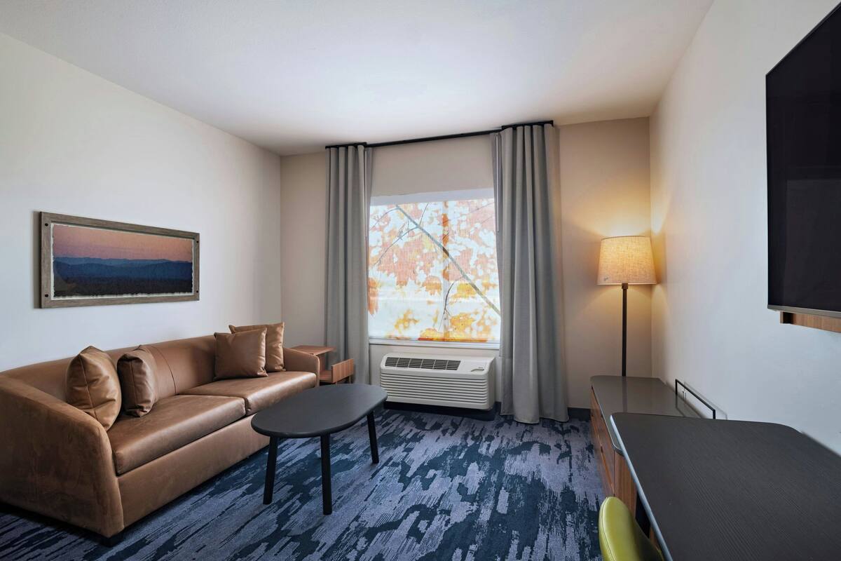 Hotellihuoneessa on ruskea nahkasohva, pieni pöytä, televisio, lamppu ja suuri ikkuna, jossa on verhot.