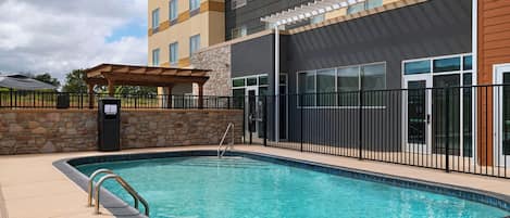 Una piscina con un muro de piedra y una pérgola, rodeada por una cerca y un edificio de varios pisos con ventanas amplias.