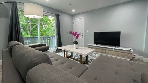 A modern living room with a grey sofa, a white coffee table, and a flat-screen TV on a wooden stand. - New, Modern 2 BR Apartment near Hospitals | HBHS (Worcester)