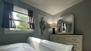 A bedroom with a bed, a dresser with drawers, a mirror, a window with curtains, and a painting on the wall. - New, Modern 2 BR Apartment near Hospitals | HBHS (Worcester)