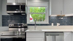 A modern kitchen with stainless steel appliances, a window with a view of trees, and a backsplash of dark tiles. - New, Modern 2 BR Apartment near Hospitals | HBHS (Worcester)