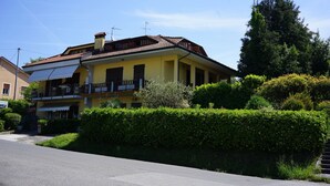 A yellow two-story house with a balcony, surrounded by greenery and a clear sky.