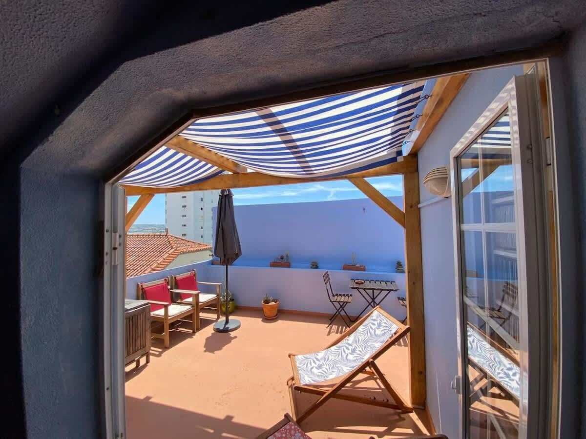 En solbelyst udendørs terrasse med en stribet markise, træmøbler og udsigt til havet.